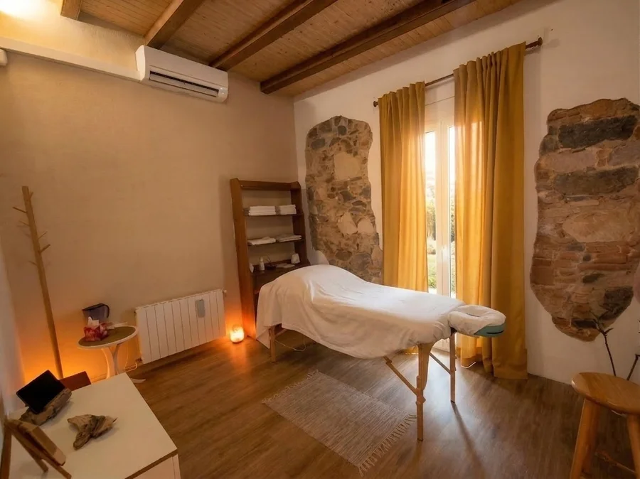 Sala de terapia corporal con camilla de masaje central, paredes blancas con secciones de piedra original expuesta, vigas de madera en el techo y cortinas mostaza por donde entra luz natural. Un espacio cálido, acogedor y consciente en Barcelona.