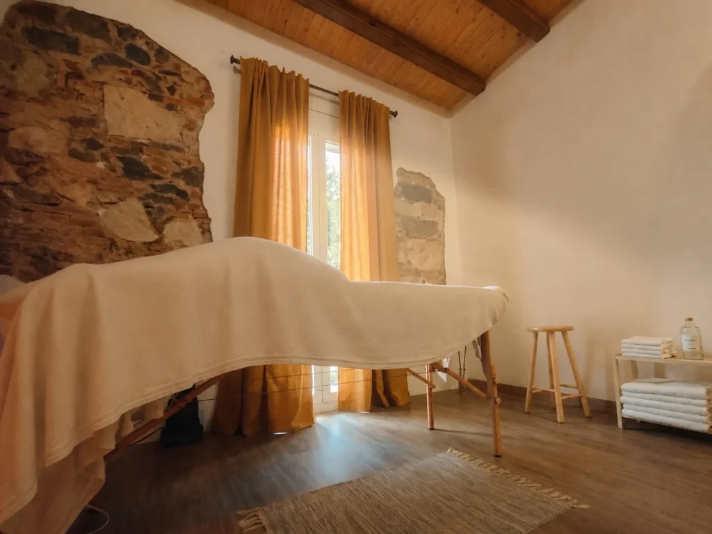 Sala de terapia corporal con camilla de masaje vestida con tejidos naturales, paredes de piedra vista antigua, vigas de madera y luz cálida filtrada por cortinas mostaza. Un espacio acogedor y con historia en Barcelona dedicado al masaje y el bienestar consciente.